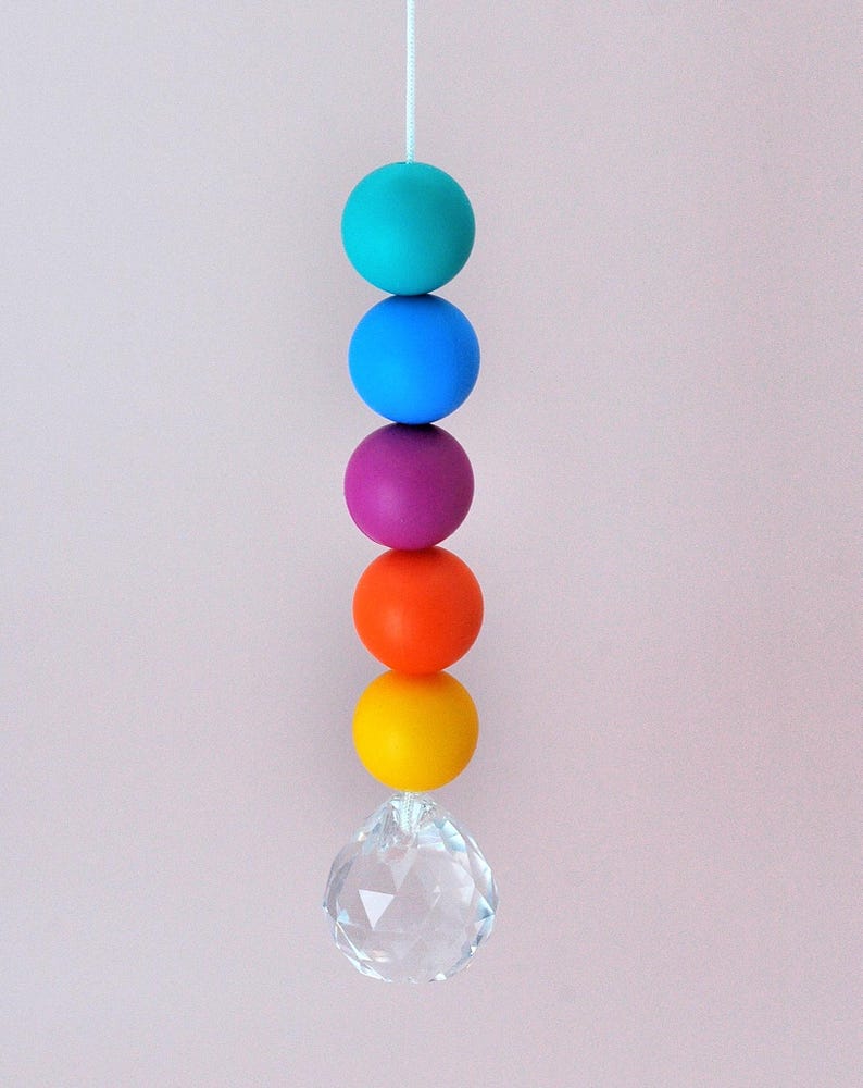 May include: A vertical hanging ornament featuring five matte spherical beads in turquoise, blue, purple, orange, and yellow, suspended from a white string. A clear, multifaceted crystal prism hangs at the bottom of the colorful bead stack.