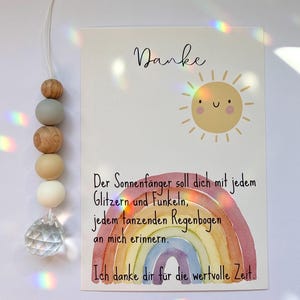 May include: A decorative suncatcher with wooden beads and a crystal prism hangs next to a card. The card features a watercolor rainbow, a smiling sun, and the word "Danke". Text in German is also visible.