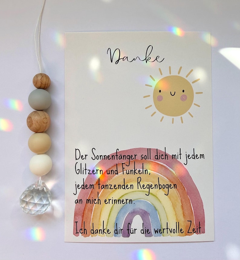 May include: A decorative suncatcher with wooden beads and a crystal prism hangs next to a card. The card features a watercolor rainbow, a smiling sun, and the word "Danke". Text in German is also visible.