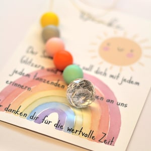 May include: A greeting card with a watercolor rainbow and a smiling sun illustration. A string of colorful beads in yellow, gray, pink, orange, and green is attached. A faceted crystal ball sits on the card. Text in German is visible.
