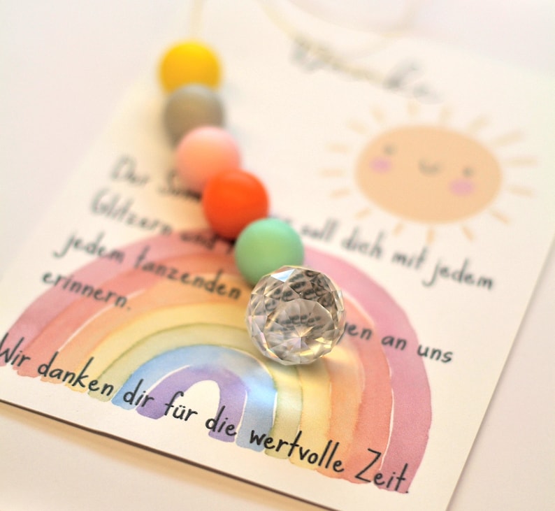 May include: A greeting card with a watercolor rainbow and a smiling sun illustration. A string of colorful beads in yellow, gray, pink, orange, and green is attached. A faceted crystal ball sits on the card. Text in German is visible.