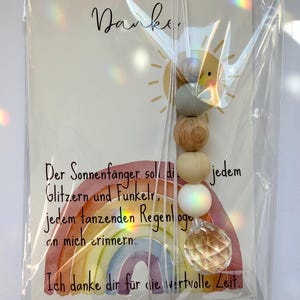 May include: A card with the word "Danke" and a sun illustration, accompanied by a rainbow-themed suncatcher. The suncatcher features wooden beads and a crystal prism, all packaged in clear plastic.