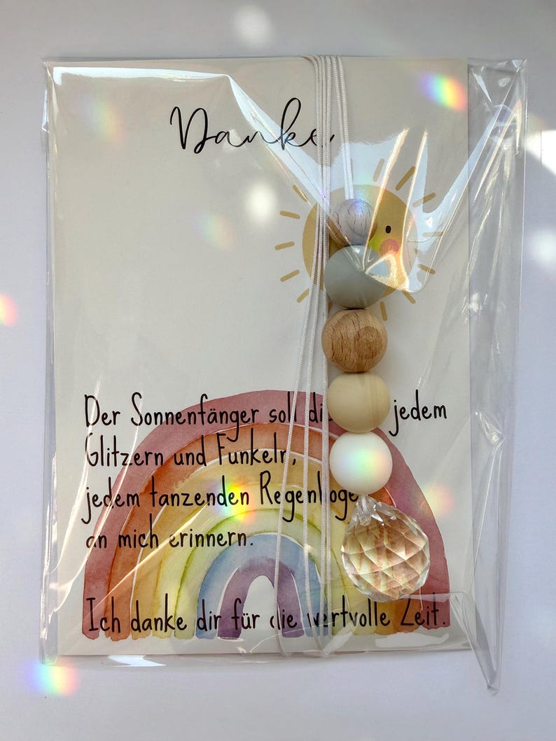 May include: A card with the word "Danke" and a sun illustration, accompanied by a rainbow-themed suncatcher. The suncatcher features wooden beads and a crystal prism, all packaged in clear plastic.