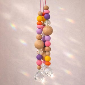 May include: A DIY suncatcher set featuring a string of colorful wooden beads in various sizes and colors, including pink, orange, yellow, and purple. The beads are strung together with a clear crystal at the bottom. Text on the image reads "Kinder lieben diesen Basteltrend".