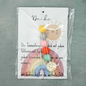 May include: A colorful suncatcher with a rainbow design and a string of beads in yellow, gray, pink, orange, and green. The card reads "Danke" and includes German text. The suncatcher is packaged in clear plastic.