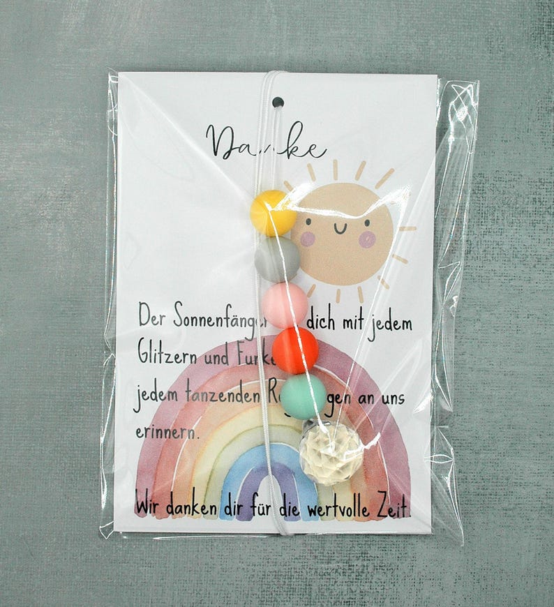 May include: A colorful suncatcher with a rainbow design and a string of beads in yellow, gray, pink, orange, and green. The card reads "Danke" and includes German text. The suncatcher is packaged in clear plastic.