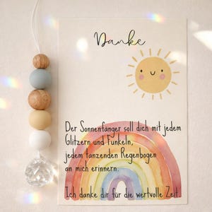 May include: A decorative suncatcher with wooden beads in shades of brown, blue, cream, and white, and a clear crystal at the bottom. A card with a watercolor rainbow, a smiling sun, and the word "Danke" is next to it.