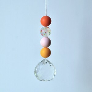 May include: A crystal prism hanging from a string with four colorful beads. The beads are orange, white with colorful sprinkles, pink, and orange.