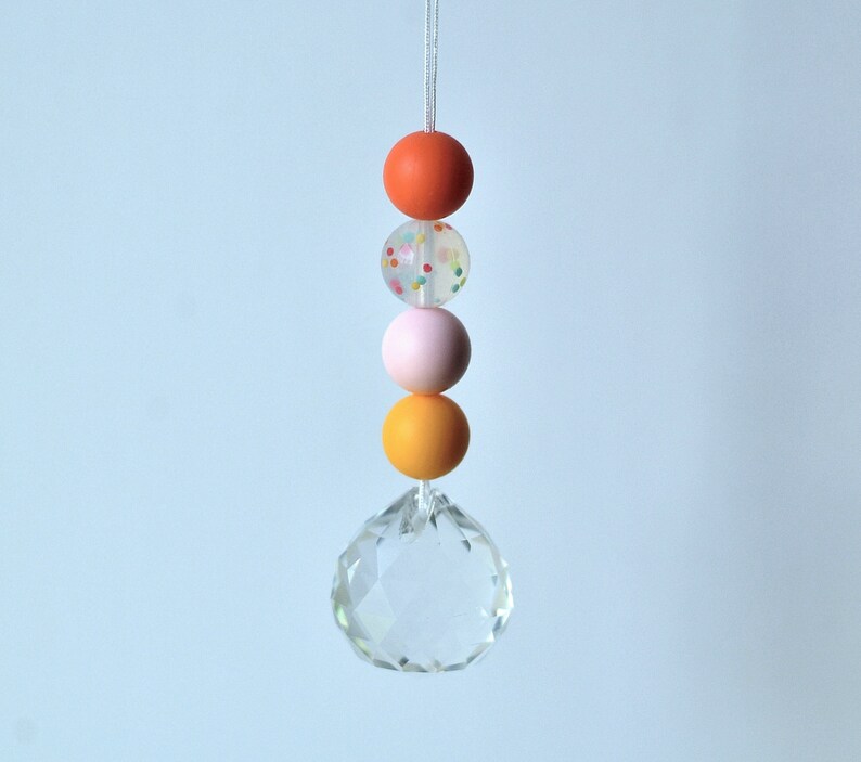 May include: A crystal prism hanging from a string with four colorful beads. The beads are orange, white with colorful sprinkles, pink, and orange.