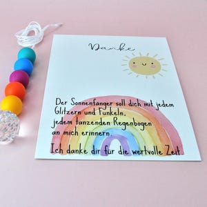 May include: A suncatcher featuring a string of colorful beads in teal, blue, purple, orange, and yellow, ending in a clear faceted crystal sphere. Beside it is a white card with a smiling sun illustration, a watercolor rainbow, and German text.