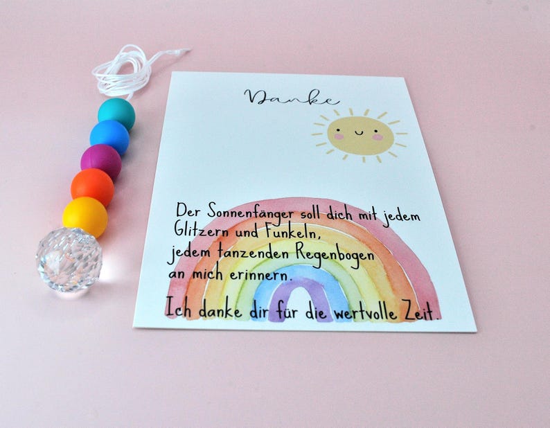 May include: A suncatcher featuring a string of colorful beads in teal, blue, purple, orange, and yellow, ending in a clear faceted crystal sphere. Beside it is a white card with a smiling sun illustration, a watercolor rainbow, and German text.