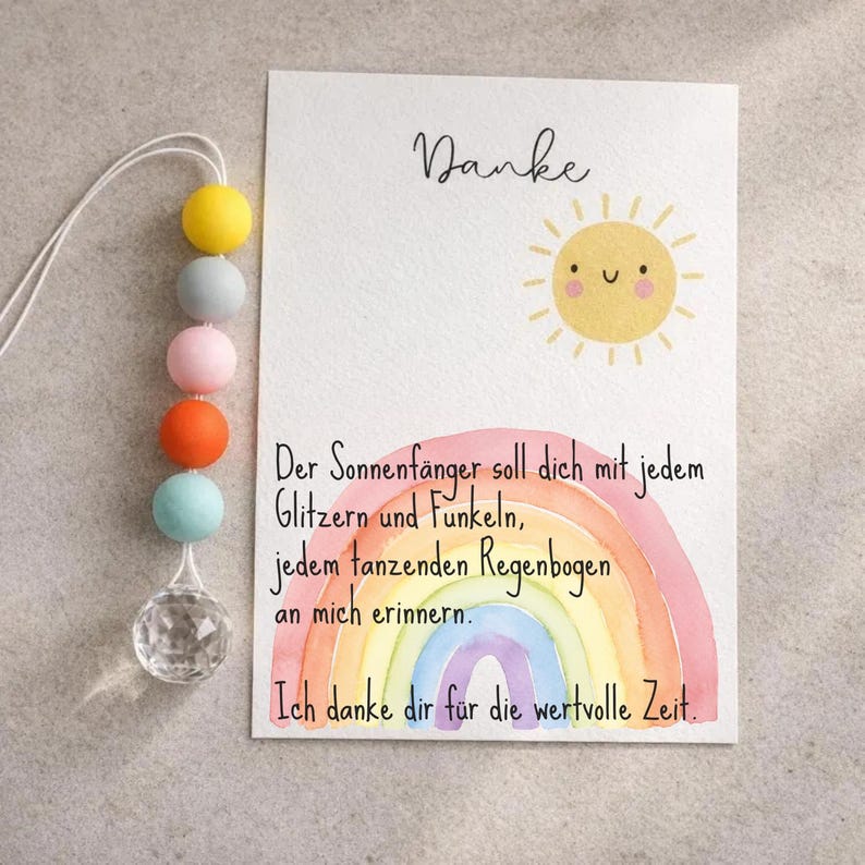 May include: A greeting card with the word "Danke" and a smiling sun illustration. The card features a watercolor rainbow and text in German. A decorative suncatcher with colorful beads and a crystal prism hangs from the top.
