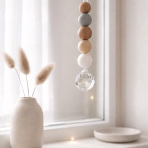 May include: A hanging decorative ornament featuring a string of wooden beads in shades of brown, gray, and white, topped with a clear crystal prism. The ornament hangs in front of a window, with a vase of dried flowers and a small dish on the windowsill.