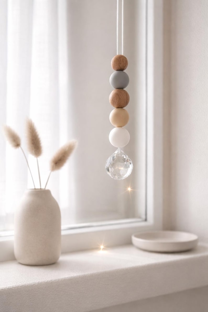 May include: A hanging decorative ornament featuring a string of wooden beads in shades of brown, gray, and white, topped with a clear crystal prism. The ornament hangs in front of a window, with a vase of dried flowers and a small dish on the windowsill.