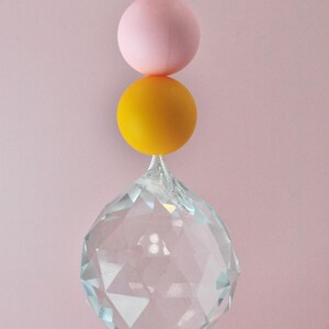 May include: A crystal prism hanging from a string with three beads: a clear bead with colorful confetti, a pink bead, and a yellow bead.