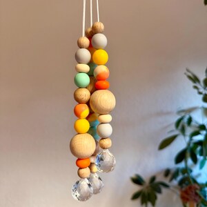 May include: A hanging mobile featuring a string of colorful wooden beads in various sizes and hues, including natural wood, gray, yellow, orange, and mint green. The mobile is accented with clear crystal pendants at the bottom, suspended by white cords.