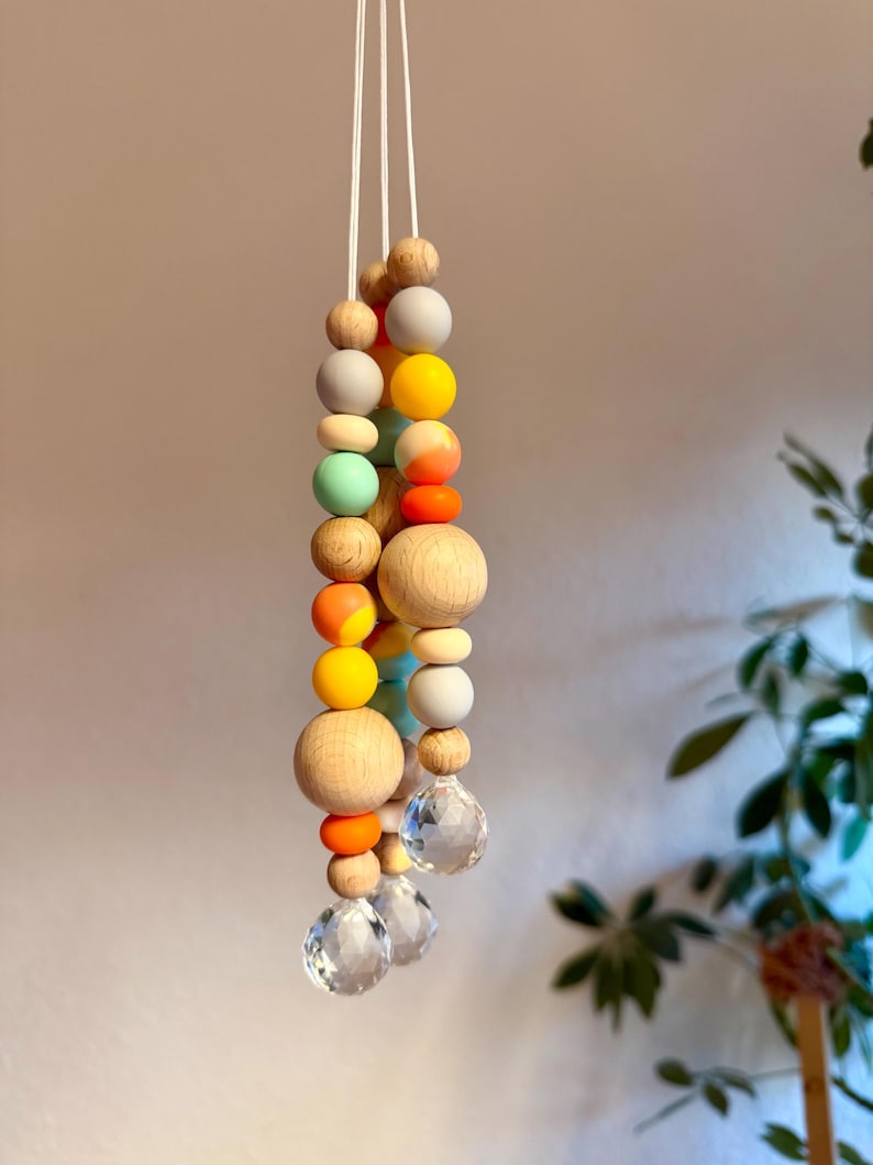 May include: A hanging mobile featuring a string of colorful wooden beads in various sizes and hues, including natural wood, gray, yellow, orange, and mint green. The mobile is accented with clear crystal pendants at the bottom, suspended by white cords.