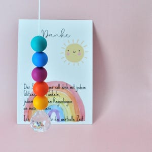 May include: A vertical string of five matte silicone beads in teal, blue, purple, orange, and yellow, ending with a clear faceted crystal pendant. Behind the beads is a white card featuring a yellow sun illustration and a watercolor rainbow.