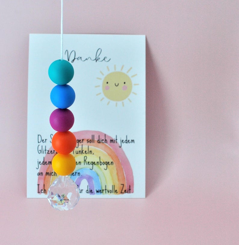May include: A vertical string of five matte silicone beads in teal, blue, purple, orange, and yellow, ending with a clear faceted crystal pendant. Behind the beads is a white card featuring a yellow sun illustration and a watercolor rainbow.