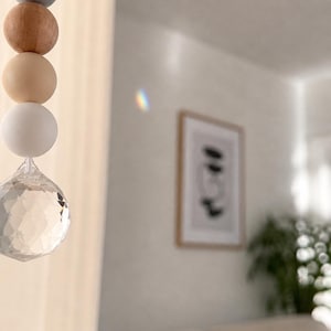 May include: A decorative hanging ornament featuring a string of wooden beads in various colors, including natural wood, gray, and cream, culminating in a faceted crystal prism. The ornament hangs in front of a blurred interior scene.
