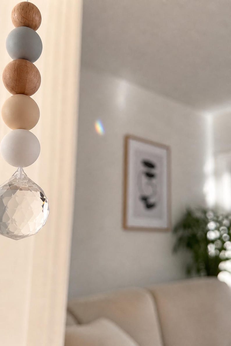 May include: A decorative hanging ornament featuring a string of wooden beads in various colors, including natural wood, gray, and cream, culminating in a faceted crystal prism. The ornament hangs in front of a blurred interior scene.