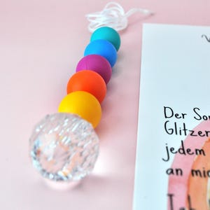May include: A vertical string of beads featuring a clear, faceted spherical bead at the bottom, followed by solid matte beads in yellow, orange, purple, blue, and teal. A white cord extends from the top bead. A white card with German text is visible.