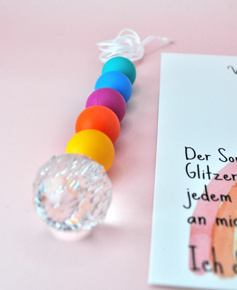 May include: A vertical string of beads featuring a clear, faceted spherical bead at the bottom, followed by solid matte beads in yellow, orange, purple, blue, and teal. A white cord extends from the top bead. A white card with German text is visible.