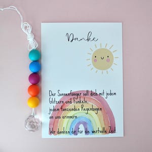 May include: A white card featuring a smiling yellow sun illustration, a watercolor rainbow, and German text that reads Danke, Der Sonnenf&auml;nger soll dich mit jedem Glitzern und Funkeln, jedem tanzenden Regenbogen an uns erinnern. Wir danken dir f&uuml;r die wertvolle Zeit. Beside the card is a string of five colorful silicone beads in teal, blue, purple, orange, and yellow, ending with a clear faceted crystal pendant.