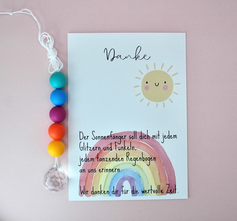 May include: A white card featuring a smiling yellow sun illustration, a watercolor rainbow, and German text that reads Danke, Der Sonnenf&auml;nger soll dich mit jedem Glitzern und Funkeln, jedem tanzenden Regenbogen an uns erinnern. Wir danken dir f&uuml;r die wertvolle Zeit. Beside the card is a string of five colorful silicone beads in teal, blue, purple, orange, and yellow, ending with a clear faceted crystal pendant.