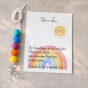 May include: A white card with the text "Danke" above a yellow sun illustration and a watercolor rainbow with German text, accompanied by a white cord featuring teal, blue, purple, orange, and yellow beads ending in a clear faceted crystal prism.