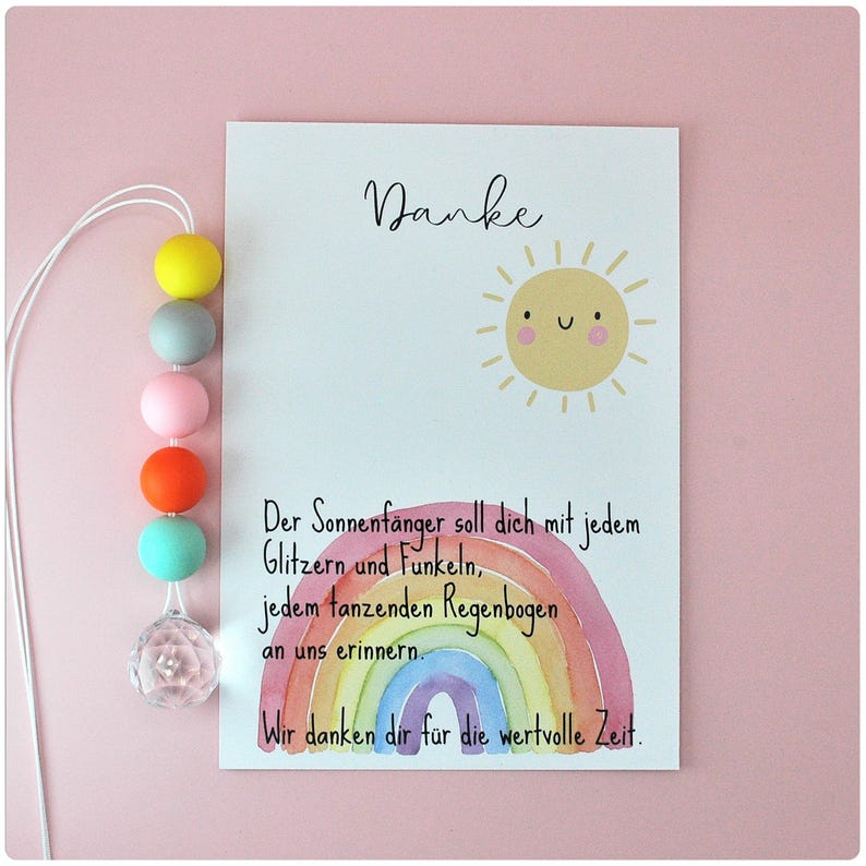 May include: A greeting card with the word "Danke" and a smiling sun illustration. Below, a watercolor rainbow and text in German. A string of colorful beads and a crystal prism hang on the left side. The background is pink.