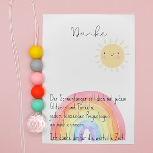 May include: A colorful beaded suncatcher with a white cord, featuring yellow, gray, pink, orange, and teal beads, and a clear crystal-like bead. A card with the word "Danke" and a rainbow illustration is next to the suncatcher.