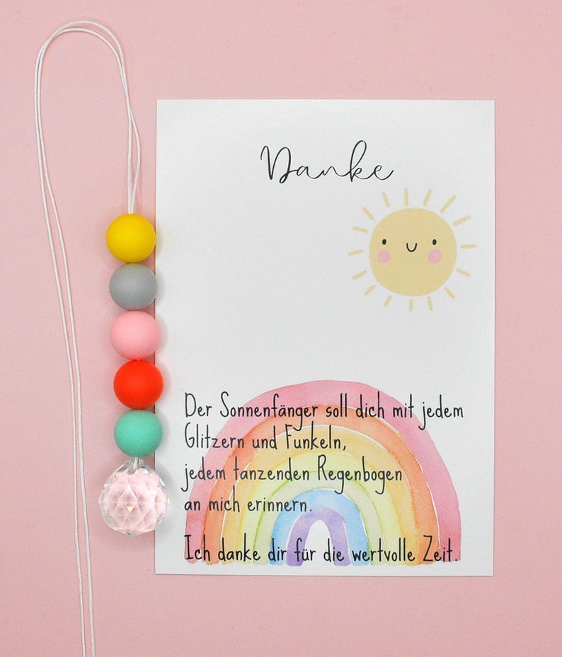 May include: A colorful beaded suncatcher with a white cord, featuring yellow, gray, pink, orange, and teal beads, and a clear crystal-like bead. A card with the word "Danke" and a rainbow illustration is next to the suncatcher.