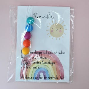 May include: A clear plastic bag containing a white card with the word Danke, a smiling sun illustration, and a watercolor rainbow. A string of five colorful beads in teal, blue, purple, orange, and yellow, plus a clear crystal charm, is attached.