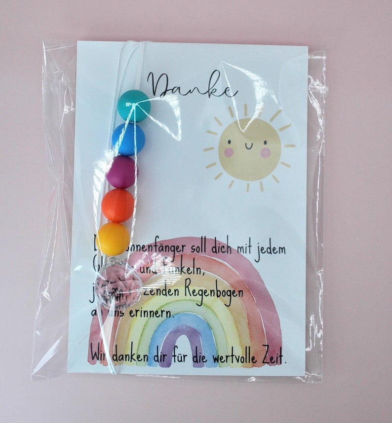 May include: A clear plastic bag containing a white card with the word Danke, a smiling sun illustration, and a watercolor rainbow. A string of five colorful beads in teal, blue, purple, orange, and yellow, plus a clear crystal charm, is attached.