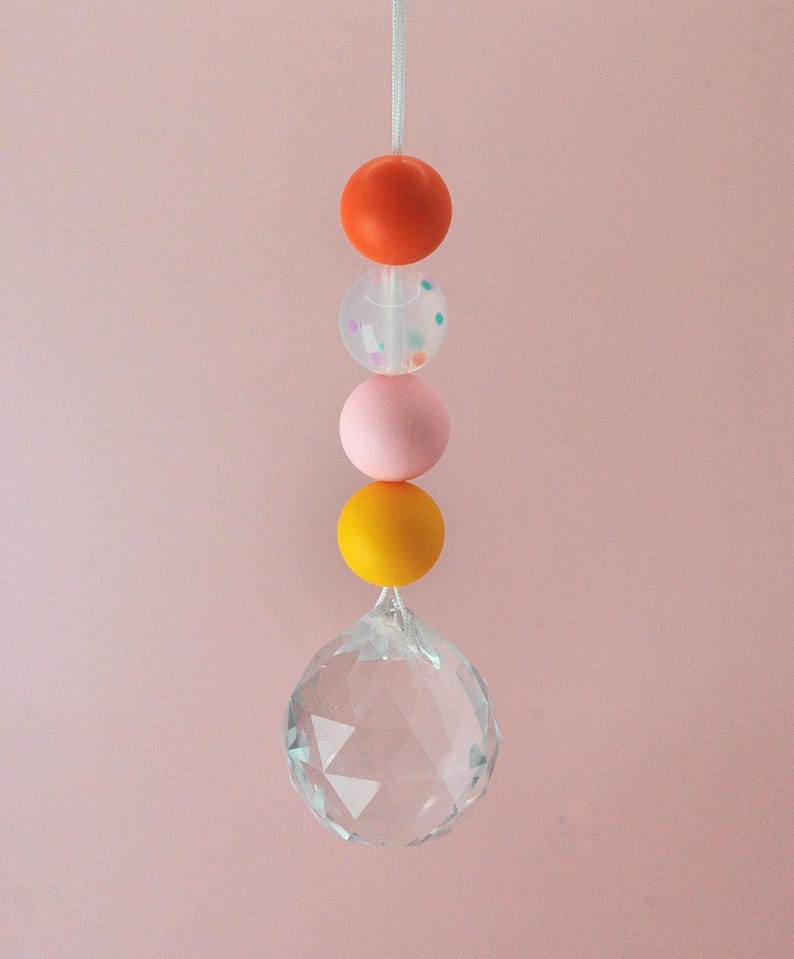 May include: A crystal suncatcher with a string of colorful beads. The beads are orange, pink, clear with confetti, and yellow.