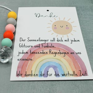 May include: A card with a watercolor rainbow and a smiling sun illustration. The card has text in German. A beaded accessory with colorful beads and a clear crystal-like charm is attached to the card.