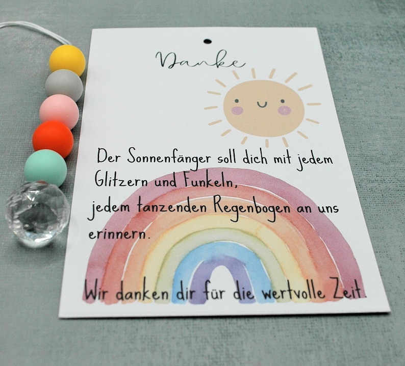May include: A card with a watercolor rainbow and a smiling sun illustration. The card has text in German. A beaded accessory with colorful beads and a clear crystal-like charm is attached to the card.