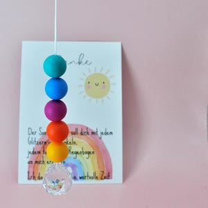 May include: A vertical string of five colorful matte silicone beads in teal, blue, purple, orange, and yellow, ending with a clear faceted crystal pendant. Behind the beads is a white card featuring a yellow sun illustration and a rainbow graphic.