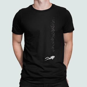 以下が含まれることがあります： 白いダイバーと泡のグラフィックが特徴の黒いTシャツ。ダイバーが上昇し、泡が上に向かって伸びています。Tシャツはクルーネックのデザインです。