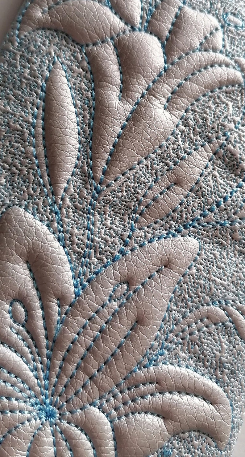 K&ouml;nnte beinhalten: Nahaufnahme einer hellgrauen Lederoberfl&auml;che mit einem blauen gestickten Blumenmuster.