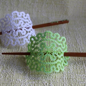 Op de afbeelding: Twee decoratieve haarstokken met witte en groene gehaakte kantversieringen. De stokken zijn gemaakt van hout en hebben een natuurlijke afwerking.