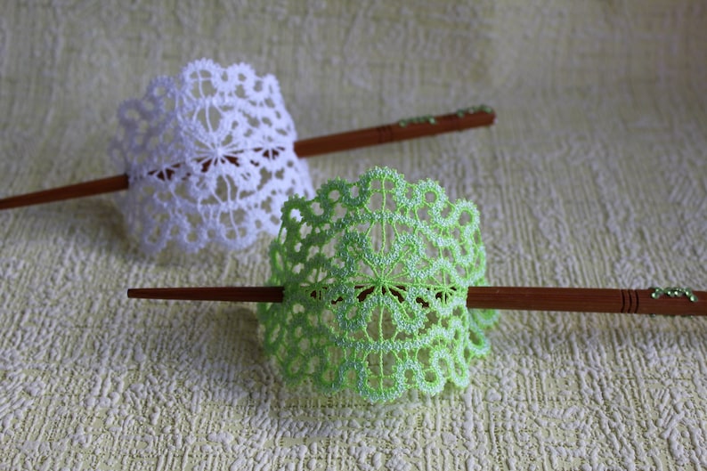 Op de afbeelding: Twee decoratieve haarstokken met witte en groene gehaakte kantversieringen. De stokken zijn gemaakt van hout en hebben een natuurlijke afwerking.