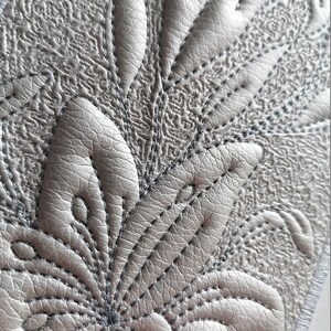 K&ouml;nnte beinhalten: Nahaufnahme eines grauen, strukturierten Stoffes mit einem dekorativen, gestickten Blumenmuster. Das Muster zeigt eine zentrale Blume mit mehreren Bl&uuml;tenbl&auml;ttern und Bl&auml;ttern.