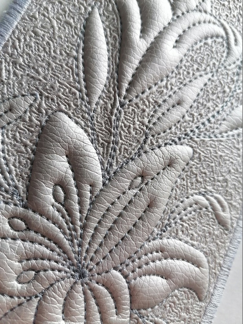 K&ouml;nnte beinhalten: Nahaufnahme eines grauen, strukturierten Stoffes mit einem dekorativen, gestickten Blumenmuster. Das Muster zeigt eine zentrale Blume mit mehreren Bl&uuml;tenbl&auml;ttern und Bl&auml;ttern.