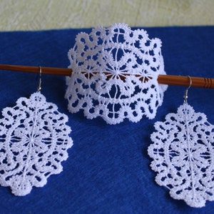 Op de afbeelding: Een set witte kant oorbellen en een bijpassende hoofdband. De hoofdband zit op een houten stok. De oorbellen zijn ovaalvormig en de hoofdband is een halve cirkel.