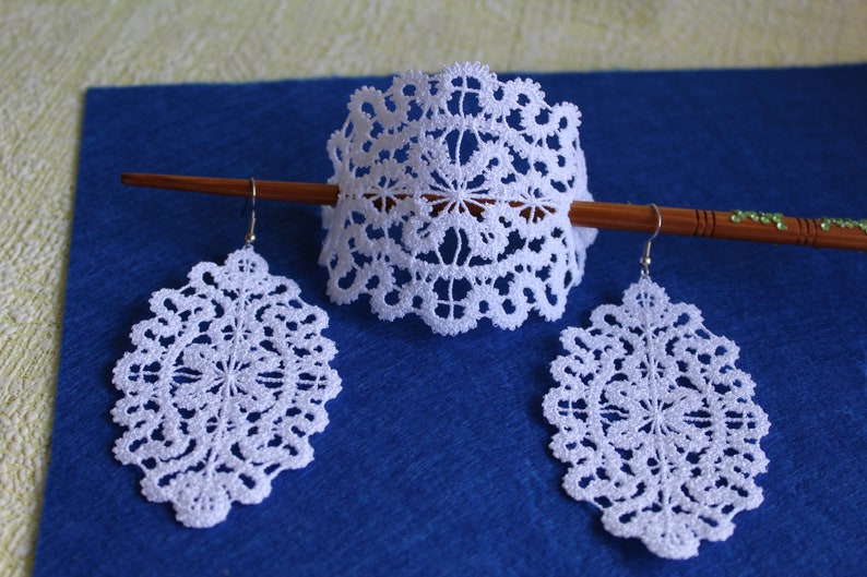 Op de afbeelding: Een set witte kant oorbellen en een bijpassende hoofdband. De hoofdband zit op een houten stok. De oorbellen zijn ovaalvormig en de hoofdband is een halve cirkel.