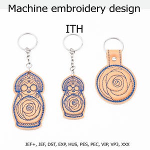 May include: Machine embroidery design ITH featuring three keychains with blue thread stitching on light tan material. Two keychains are shaped like nesting dolls with glasses and rose motifs, and one is circular with a rose design. JEF+, JEF, DST, EXP, HUS, PES, PEC, VIP, VP3, XXX.