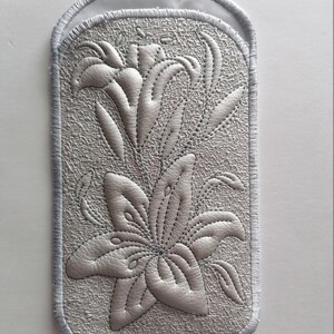 K&ouml;nnte beinhalten: Ein wei&szlig;er gestickter Aufn&auml;her mit einem Design aus zwei Lilien. Der Aufn&auml;her hat einen wei&szlig;en Satinrand.