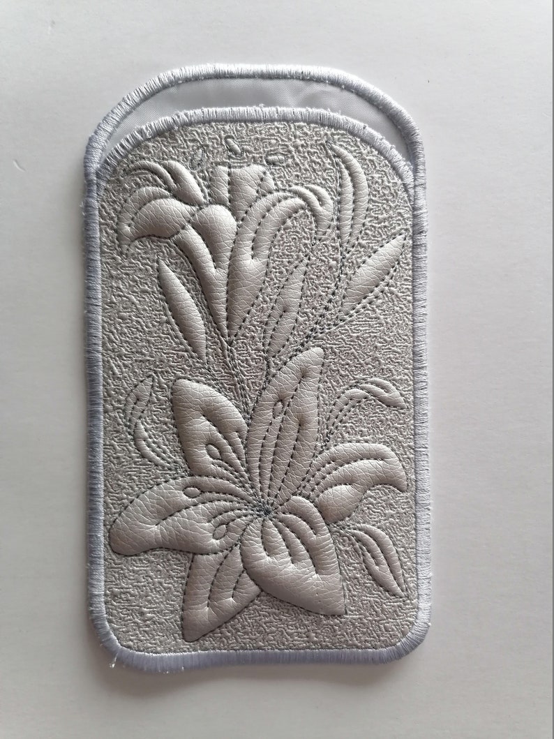 K&ouml;nnte beinhalten: Ein wei&szlig;er gestickter Aufn&auml;her mit einem Design aus zwei Lilien. Der Aufn&auml;her hat einen wei&szlig;en Satinrand.